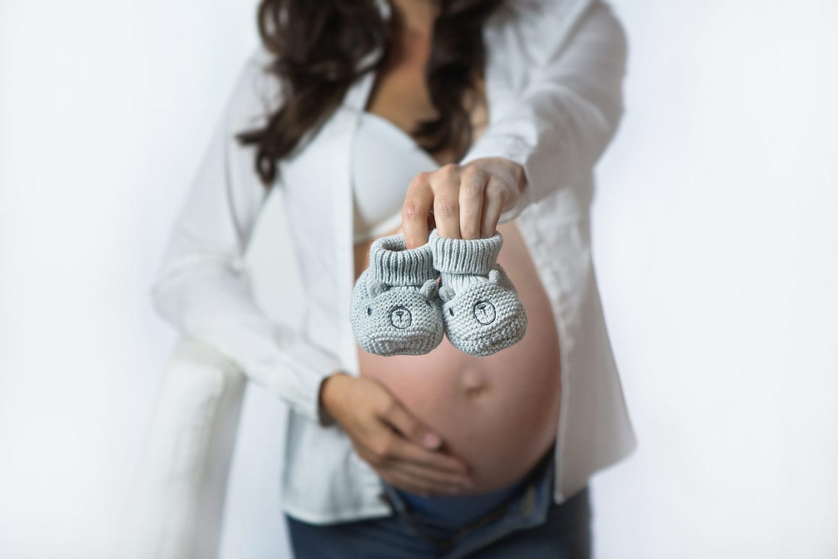 Die schwangere Dame zeigt dem Babybauch und den kleinen Schuhen in ein Babybauch Fotoshooting von Donna Bellini Berlin