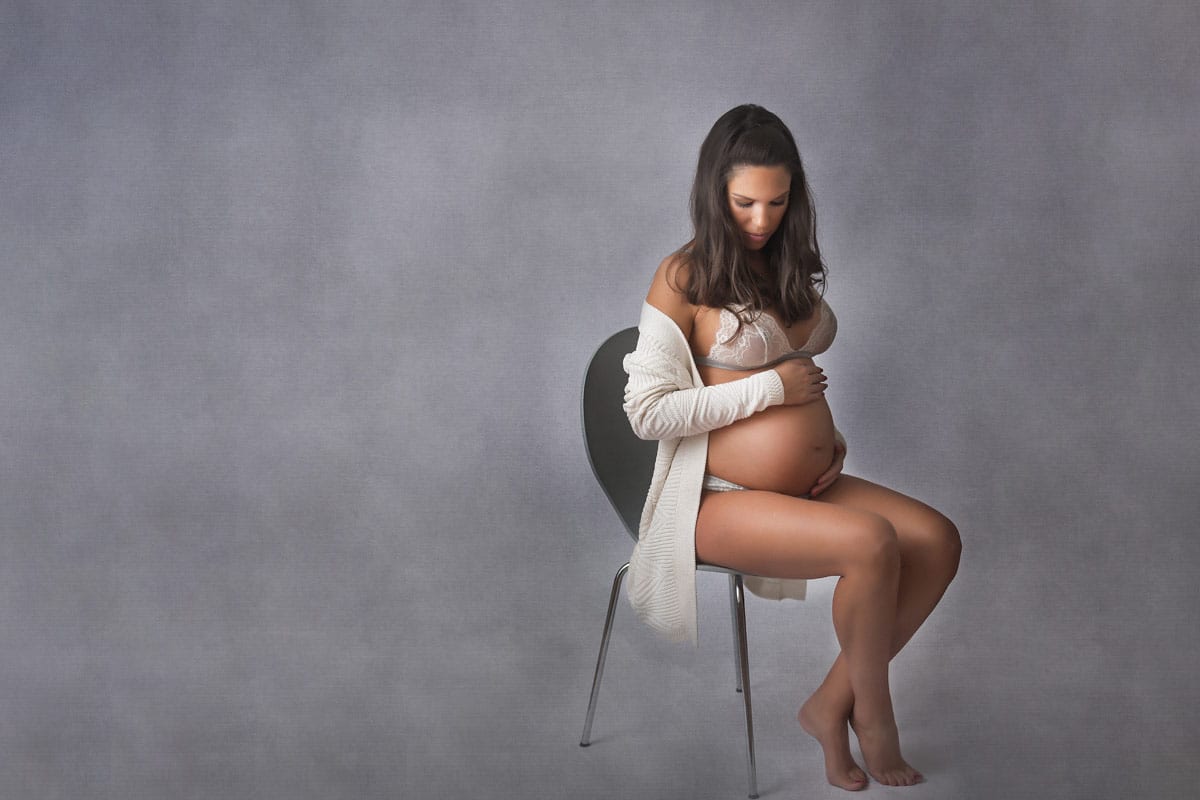 Schwangere Frau sitzt und posiert auf grauem Hintergrund ein Schwangerschaftsfotoshooting von Donna Bellini Berlin
